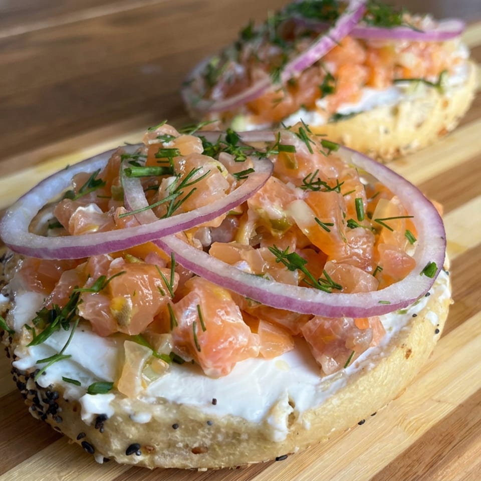 Open-faced bagels topped with creamy spread, diced salmon, red onion rings, and fresh herbs on a wooden serving board.