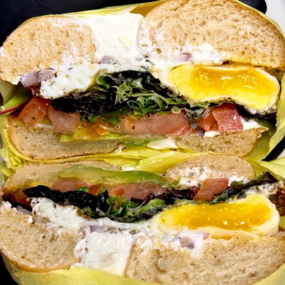 Two loaded sandwiches with fried eggs, leafy greens, tomatoes, pickles, and creamy sauce, held in gloved hands.