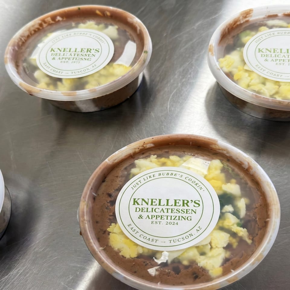 Lidded containers of Kneller’s Delicatessen and Appetizing food arranged on a stainless steel countertop.